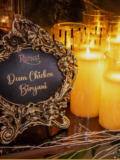 An ornate, vintage-style frame used as a food label for our Dum Chicken Biryani, surrounded by the warm glow of candles. Every detail is an opportunity for elegance.