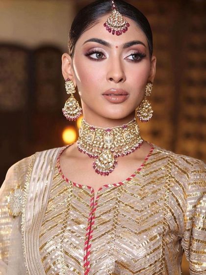 A close-up of a classic bridal look with a modern twist. The makeup features a sharp, defined eye and a soft nude lip, perfectly complementing the gold and ruby jewellery.