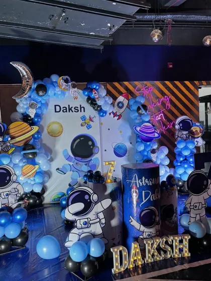 An out-of-this-world space theme birthday for a 7-year-old. This detailed setup includes astronaut and planet cutouts, a galaxy backdrop, marquee letters, and a blue and black balloon arrangement to create a cosmic adventure.