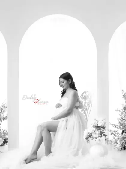 A dreamy, ethereal portrait in black and white. The mom-to-be is styled with angel wings and seated on a bed of clouds, creating a heavenly and serene image.