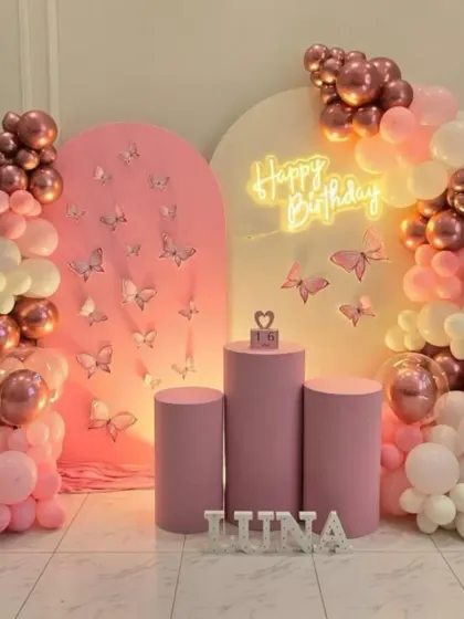 A modern and chic butterfly-themed birthday party. The setup includes two pink arched backdrops, one with a textured design, and a beautiful balloon arrangement in white, pink, and rose gold.