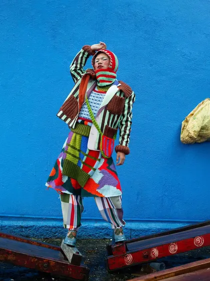 A powerful stance against a bold blue wall. This image from the "Kaleidoscope" series highlights the full outfit, showcasing the complex layering of patterns and textures that define this avant-garde collection.