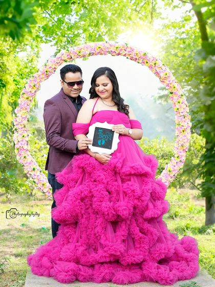 A happy couple holding a sonogram picture, framed by a circular floral prop. The mother-to-be's vibrant pink gown is a showstopper.