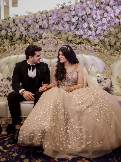 A fairytale moment from an engagement ceremony. The couple is seated on an elegant sofa against a stunning wall of white flowers, creating a truly grand and romantic scene.