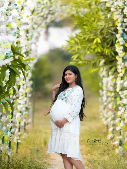 A beautiful outdoor portrait of a mother-to-be in a simple white dress, standing under a floral arch. The natural light and serene setting create a fresh and timeless image.