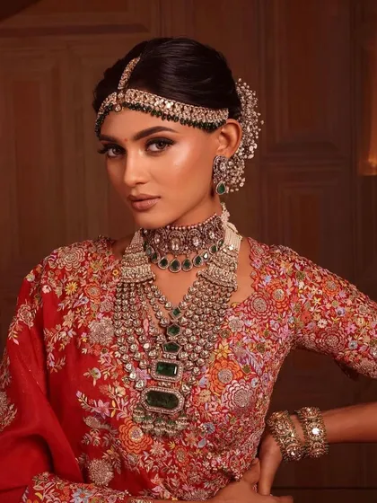 A close-up of a regal bridal look. The makeup is warm and defined, with a focus on the eyes, complementing the rich red and gold lehenga and the intricate jewellery.