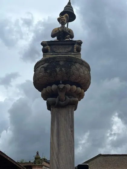 A stone pillar in Bhaktapur Durbar Square, standing tall against a dramatic sky. The city's atmosphere is thick with history and reverence.