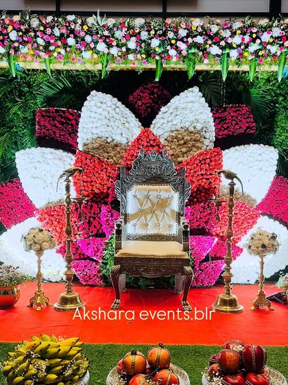 A stunning Seemantham stage featuring a large floral lotus made of red, white, and gold-colored flowers. The backdrop is a lush green wall, and the stage is set with traditional offerings.
