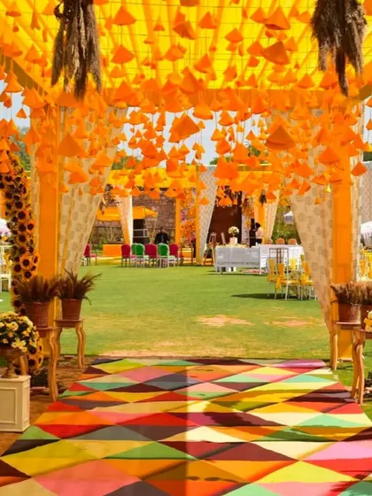 A colorful and festive entrance for a Haldi carnival. The walkway is a vibrant multi-color carpet, leading into a space decorated with hanging orange cones and marigold arches.