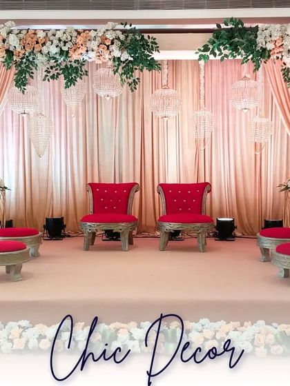 A full view of the chic mandap stage, showcasing the symmetrical arrangement of red chairs, pastel drapes, and a canopy of beautiful white and peach flowers.