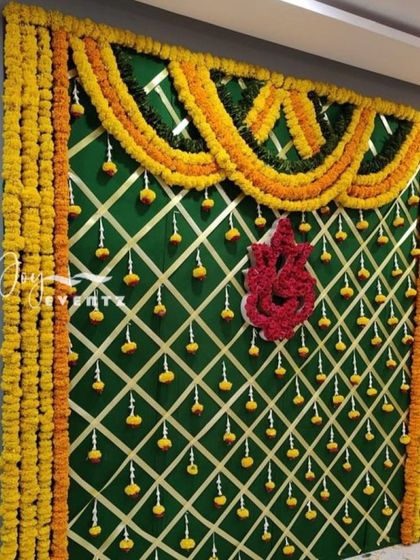 This traditional half saree function backdrop features a Ganesha motif made of red flowers against a green fabric background. The design is framed with marigold garlands and accented with a crisscross ribbon pattern.