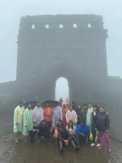 A group photo in front of the massive gate of a fort in Maharashtra, completely enveloped in fog and rain.