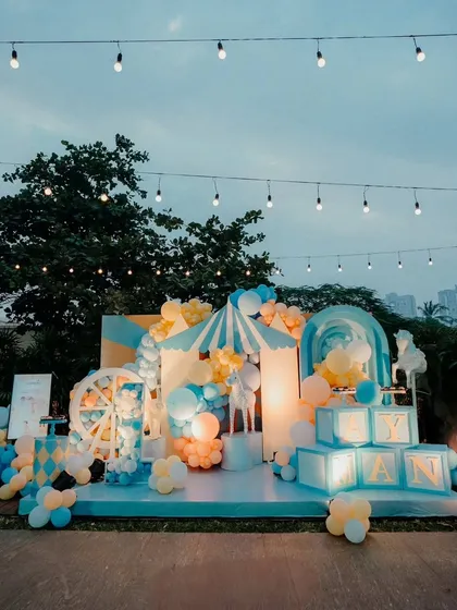 A vibrant and playful carnival-themed first birthday party. This outdoor setup includes a custom-built stage with a Ferris wheel prop, a circus tent backdrop, and light-up name blocks, all in a cheerful blue and yellow palette.