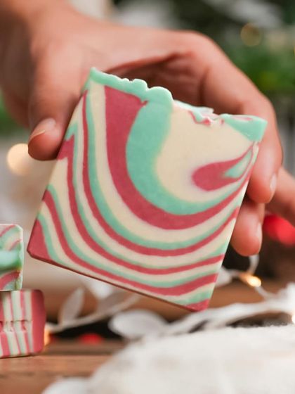 A hand holding a beautifully swirled red, green, and white soap bar. This shot highlights the artisanal, handcrafted quality of the product.