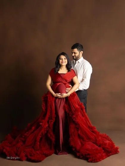 A classic portrait of a couple against a rich brown studio backdrop. The mom-to-be is featured in a dramatic red ruffled gown, standing with her partner.