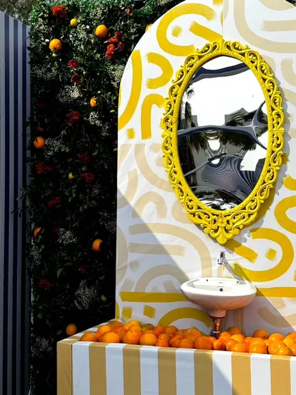 A detailed shot of the unique vanity setup. The sink is filled with fresh oranges, and the ornate yellow mirror provides a perfect spot for selfies against the artistic backdrop.