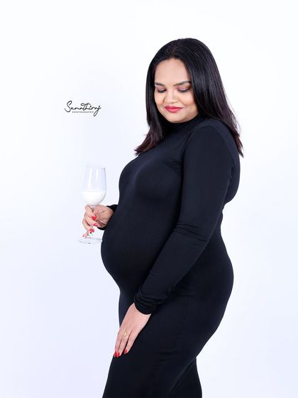 A simple and classic portrait of a mother-to-be in a black dress, holding a glass of milk. The clean white background keeps the focus on her radiant glow and elegant posture.
