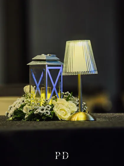 A simple yet elegant centerpiece for a cocktail event, featuring a modern lamp, a classic lantern, and a small arrangement of white roses.