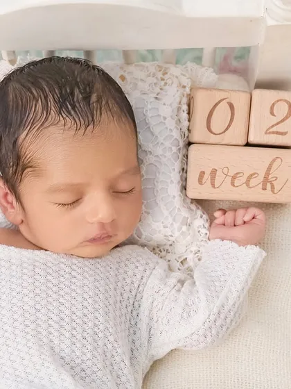 A close-up portrait of a two-week-old newborn, with wooden blocks marking their age. The focus is on their peaceful expression and the soft textures surrounding them.