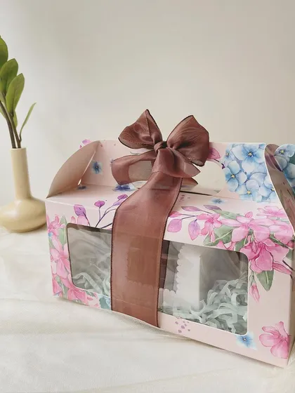 The Rakhi Celebration Hamper, presented in a beautiful floral box with a brown ribbon. This gift set includes our popular choco almonds, wheat double chocolate cookies, rich brownies, and our signature Regal Riches.