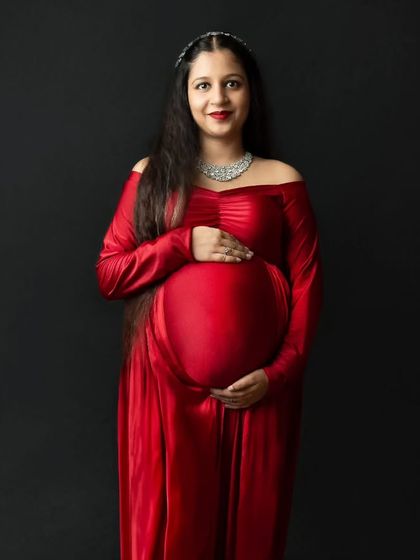 A classic and direct portrait of a radiant mother-to-be in a striking red gown against a solid black background. This simple setup puts all the focus on her joyful expression and beautiful baby bump.