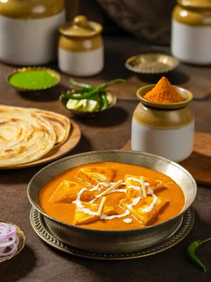 Shahi Paneer in a traditional metal bowl, surrounded by pickling jars and parathas. This composition for Rakesh Masala creates a feeling of a complete, wholesome meal.
