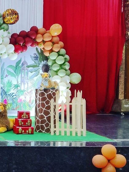 A section of a jungle-themed birthday decoration showing a giraffe-print plinth, a stuffed lion, and a colorful balloon arch against a red curtain backdrop.