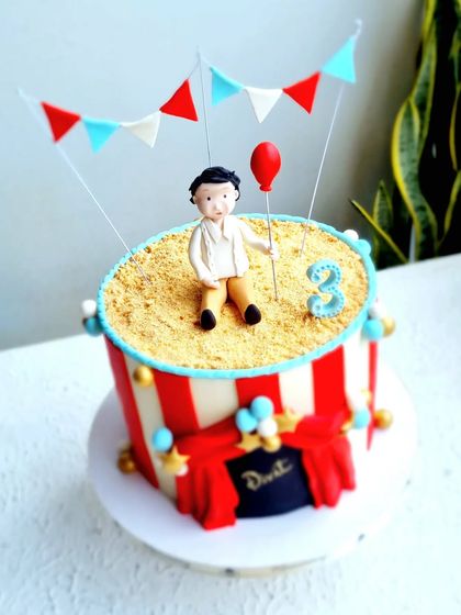 A carnival-themed cake with a figurine sitting on a "sand" top made of crushed biscuits.