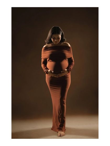 A portrait of quiet contemplation. The mother-to-be looks down at her belly with a gentle expression, her hands cradling her bump, in this beautifully lit, full-length studio shot.