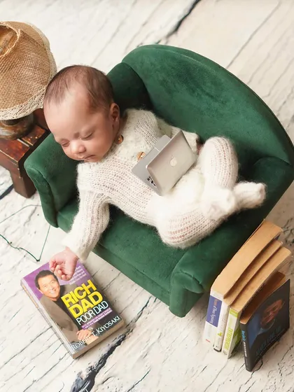 CEO of midday naps! This humorous setup features a newborn sleeping on a miniature couch with a tiny laptop and a copy of 'Rich Dad Poor Dad'.