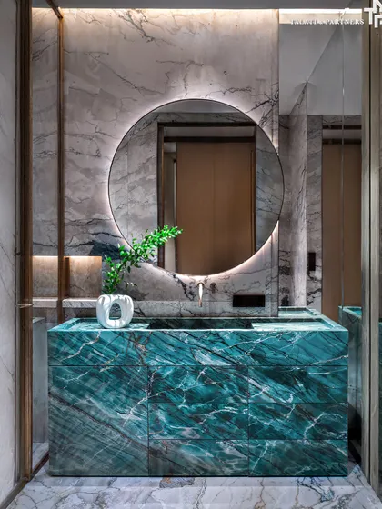 A bold bathroom design featuring a custom vanity crafted from a dramatic green marble. Set against a lighter marble wall and floor, the vanity becomes a sculptural centerpiece, complemented by a large, backlit circular mirror.