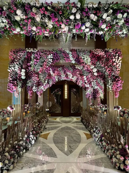 A straight-on view of the magnificent floral entrance. The archway, thick with purple and white flowers, perfectly frames the doorway, inviting guests into a world of celebration.
