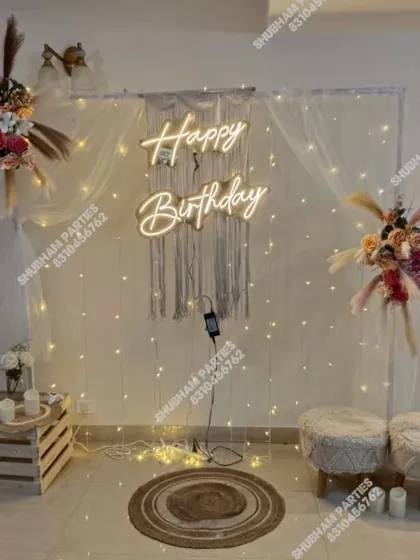 A stylish and modern boho-themed birthday decoration perfect for an intimate celebration. This setup features a macrame wall hanging, a "Happy Birthday" neon sign, fairy lights, and dried floral arrangements for a chic and rustic vibe.