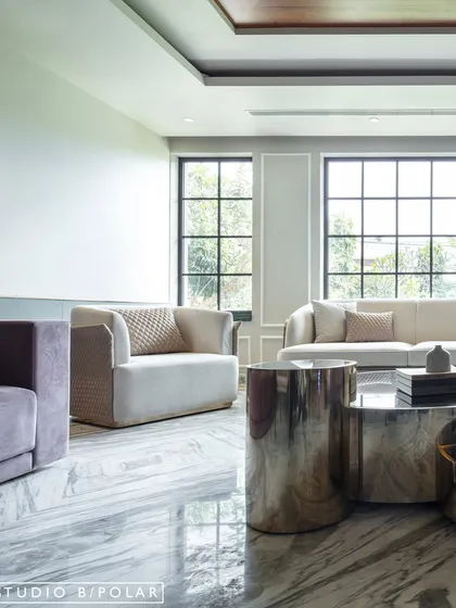 The seating arrangement includes sofas and armchairs in soft, neutral tones with quilted details, creating a comfortable yet sophisticated atmosphere. Large windows flood the room with natural light.