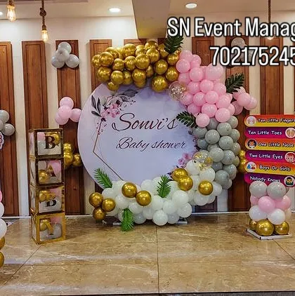 Here is an elegant baby shower setup for a party hall. It features a personalized circular backdrop with the mother's name, surrounded by a custom balloon arrangement in pink, gold, and sage green. I can match any color scheme you have in mind.