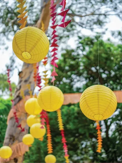 Floating yellow paper lanterns and colorful hanging garlands create a dreamy, whimsical ambiance for an outdoor Mehendi. They add a touch of playfulness and look beautiful against the natural greenery.