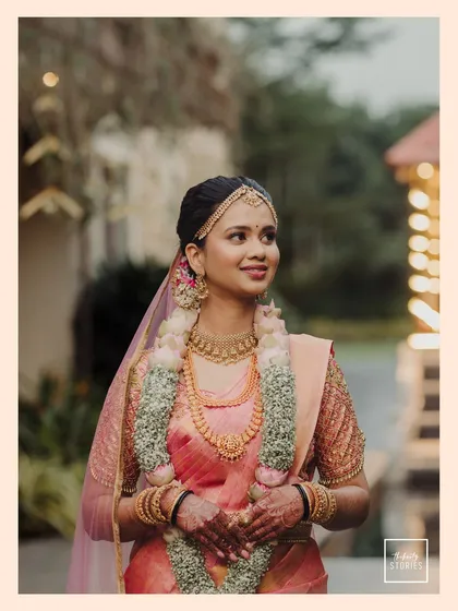 A classic bridal portrait taken outdoors. The soft, natural light beautifully illuminates the bride in her peach-colored saree and traditional jewelry.