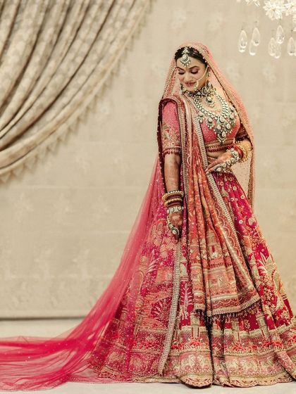 A full-length portrait of the bride in a magnificent pink and red lehenga, its long train spread out, showcasing the grandeur of her bridal look.