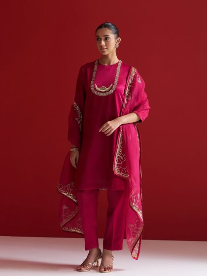 A model stands against a bold red background in a magenta kurta set, featuring a beautifully embroidered dupatta draped elegantly over her shoulder.