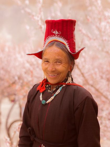 A smiling portrait of a woman from Ladakh, her face weathered by the high-altitude sun. Her traditional red hat and turquoise necklace highlight her cultural identity.