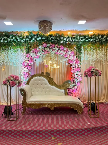 For an elegant engagement, anniversary, or pre-wedding party, we design stunning stage setups. This one features a beautiful floral ring with a 'Better Together' neon sign, complemented by a classic chaise lounge and matching flower stands, creating a sophisticated focal point in a banquet hall.