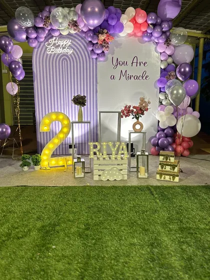 A lovely lavender-themed second birthday decoration. The setup features a double arch backdrop and a purple balloon garland, complemented by a large marquee number '2' and personalized name display.