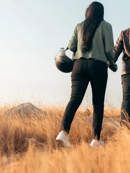 Walking into the future together. Carrying their helmets, the couple walks hand-in-hand through a field of tall, golden grass, ready for the next leg of their adventure.