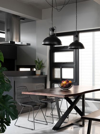 The dining area blends industrial edge with natural touches. The dark metal pendant lights and wireframe chairs are softened by the live-edge wood table and indoor plants, creating a balanced and inviting space for meals.