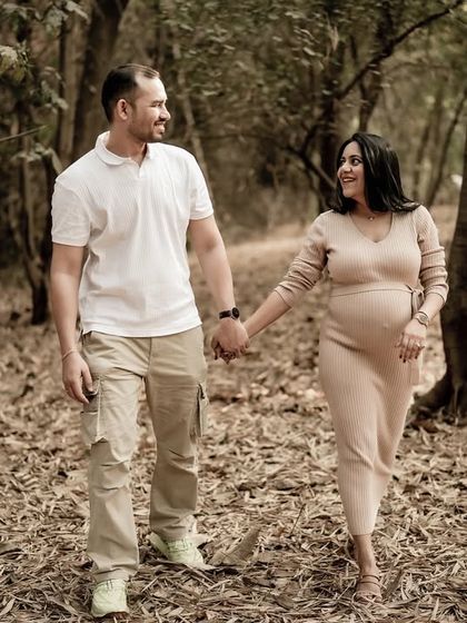 The couple walks hand-in-hand through a path in the woods, sharing a smile. The natural setting creates a peaceful and intimate atmosphere.