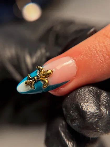 A close-up of a 3D fleur-de-lis charm on a blue and white abstract nail. Adding a single, bold charm like this can completely elevate a nail set.