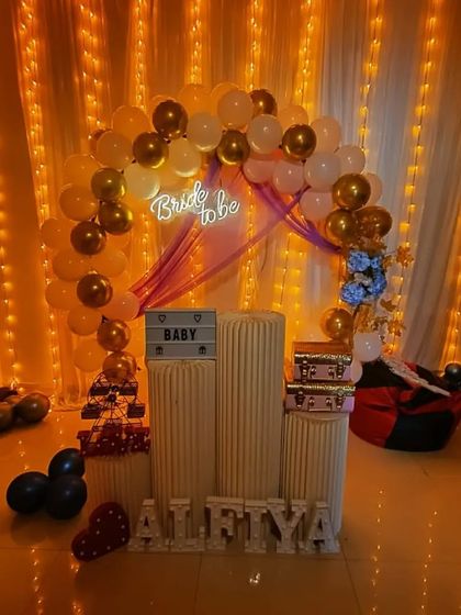 An elegant bride-to-be or baby shower setup with a gold and white balloon arch. The decor includes a "Bride to be" neon sign and personalized light-up letters.