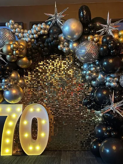 A dazzling 70th birthday setup. We used a gold sequin shimmer wall as the base, framed by a black, silver, and gold balloon arch with starburst accents and large light-up numbers.