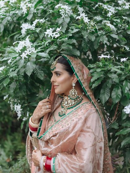 A beautiful portrait of a Sikh bride in her pastel pink and green attire, standing gracefully amidst lush greenery.