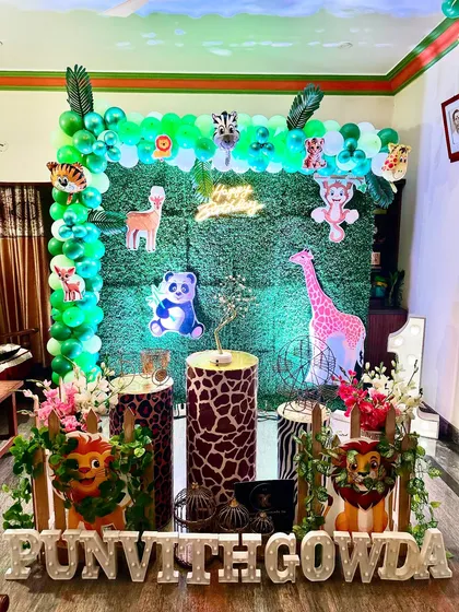 A jungle-themed decoration perfect for a home setup. It features a green hedge backdrop adorned with animal cutouts, animal-print plinths, and marquee letters for the birthday boy's name.